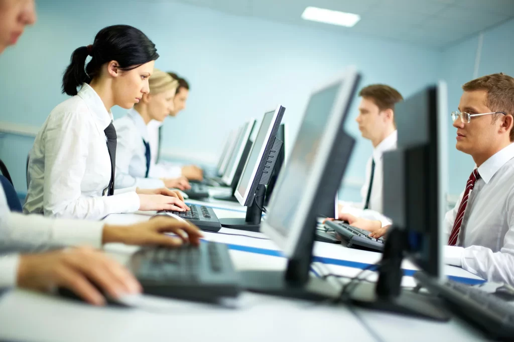 Mesa com vários computadores e pessoas trabalhando neles.